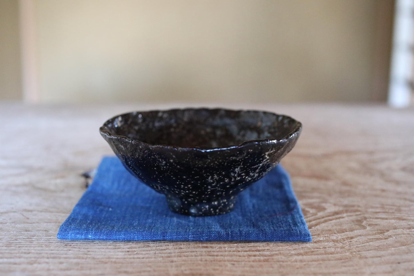 A black ceramic bowl with a speckled texture sits on a blue cloth on a wooden surface.