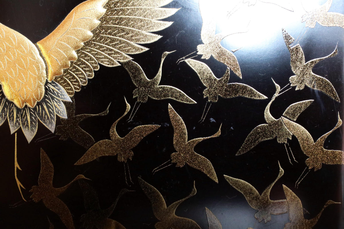 a close-up view of a black background with a pattern of gold-colored cranes. The cranes are depicted in various sizes and positions, creating a dynamic and visually striking composition.
