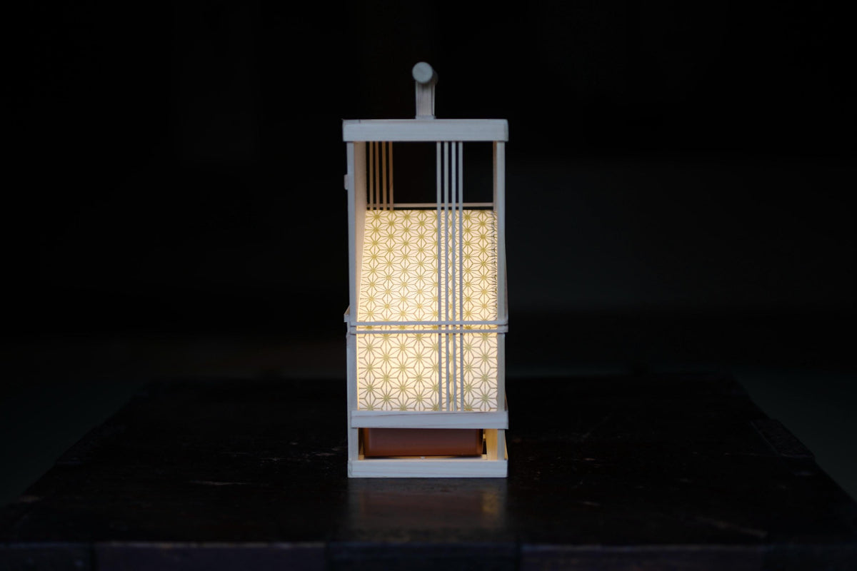 A white lantern with a patterned design is lit, casting a warm glow on a dark surface.