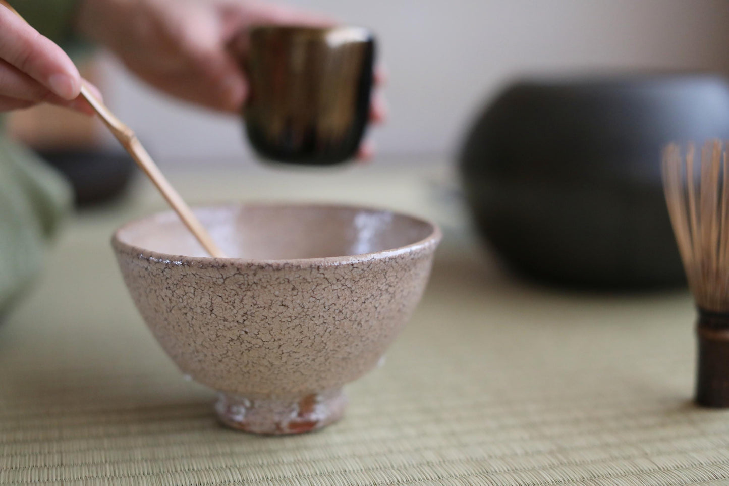 A hand is holding a small bowl with a wooden spoon, and there is a cup of tea in the background.