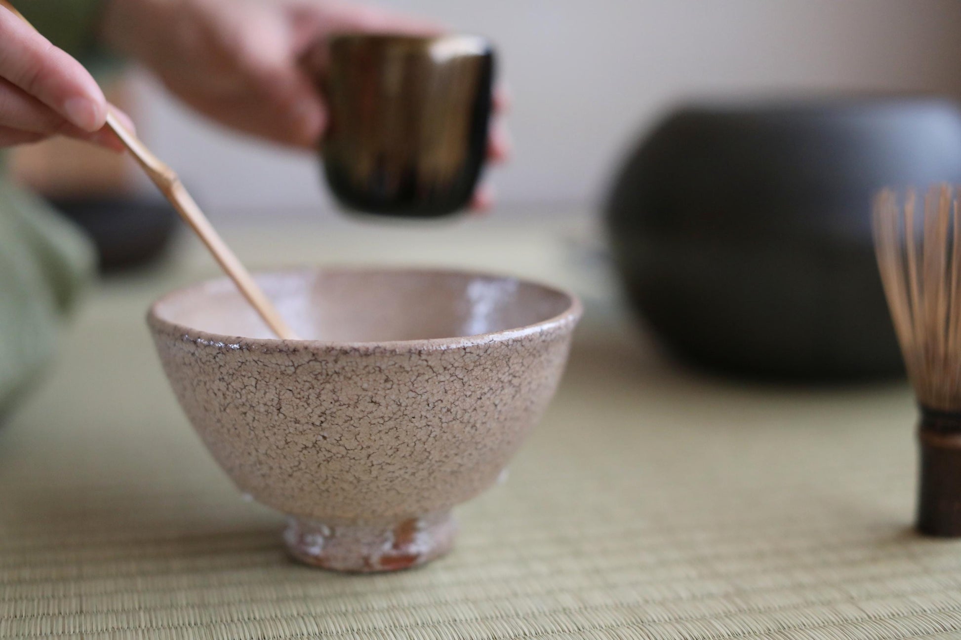A hand is holding a small bowl with a wooden spoon, and there is a cup of tea in the background.