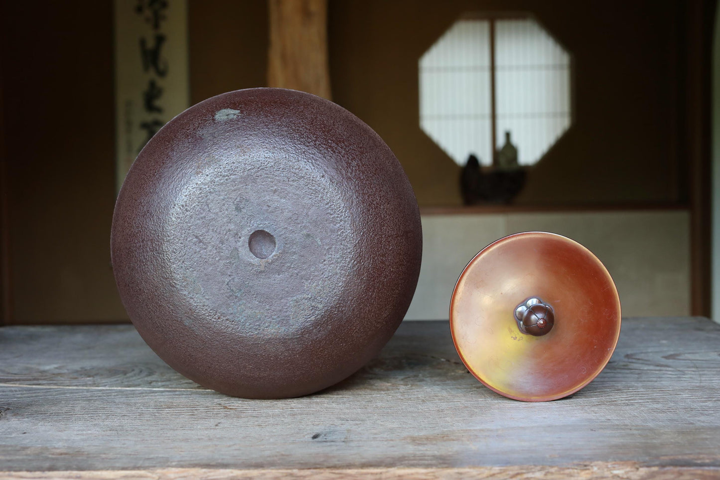 two ceramic objects placed on a wooden surface. One object is a large, round, brown ceramic piece, while the other is a smaller, round ceramic piece with a copper-like finish.