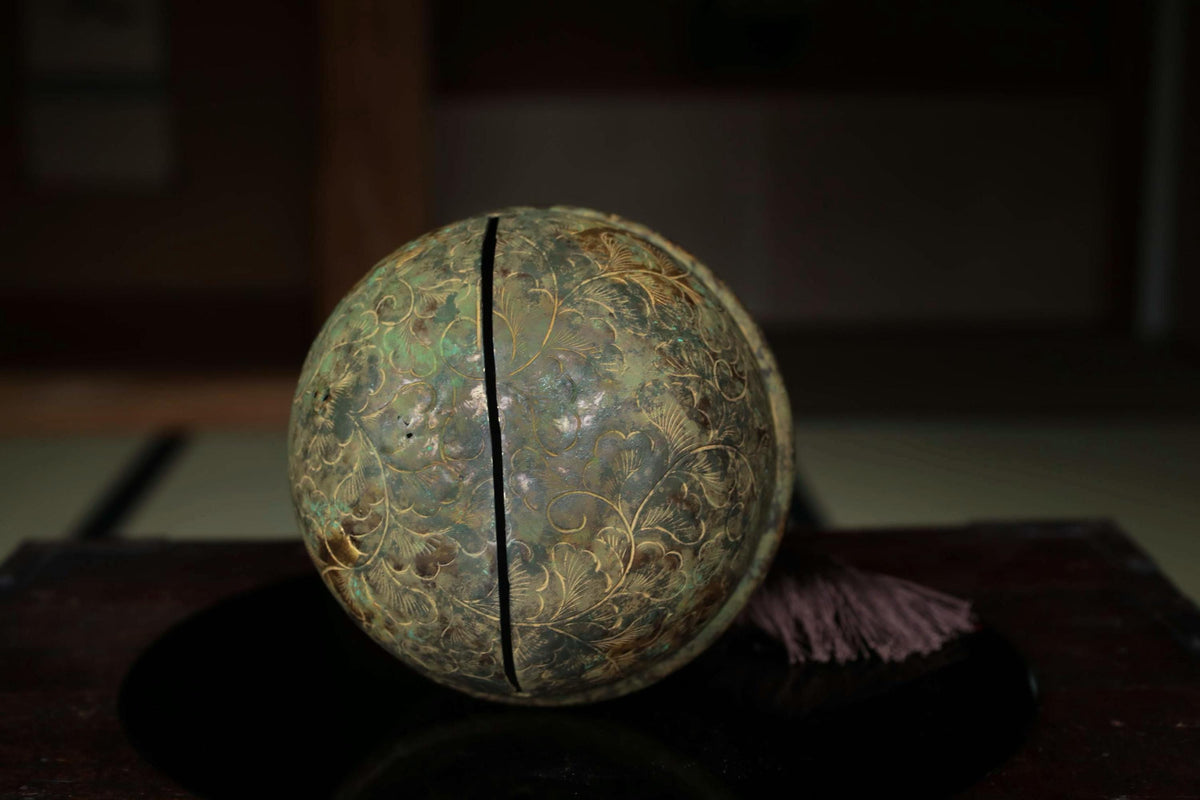 An old, ornate metal ball with a floral pattern sits on a dark surface, with a small purple object next to it.