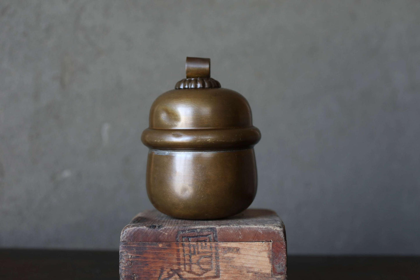 A brass container or jar with a round lid sits on a wooden surface against a gray background.