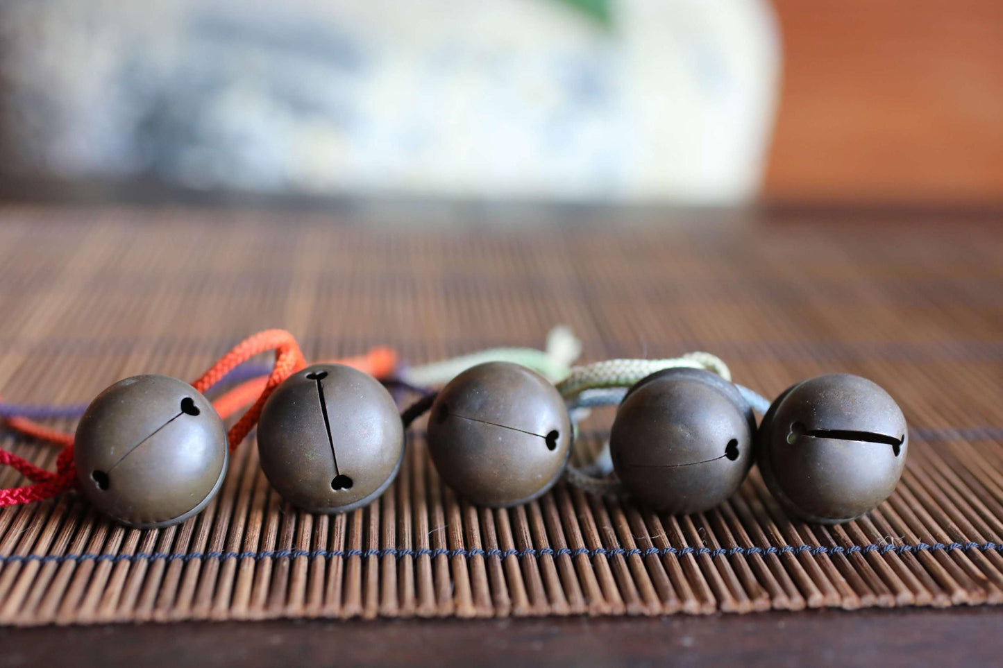 A row of five metal bells with different colors, including red, orange, and blue, are arranged on a bamboo mat.