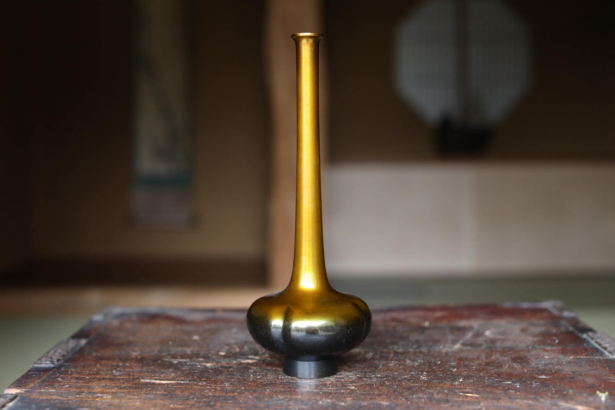 A golden vase with a long, slender shape sits on a wooden surface, with a blurred background suggesting an indoor setting.