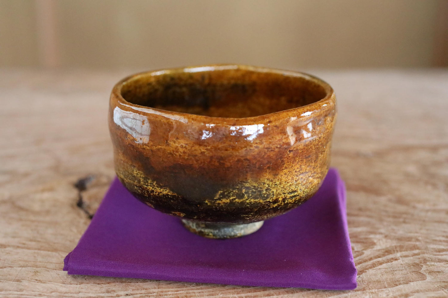 A ceramic bowl with a brown and orange glaze sits on a purple cloth on a wooden surface.