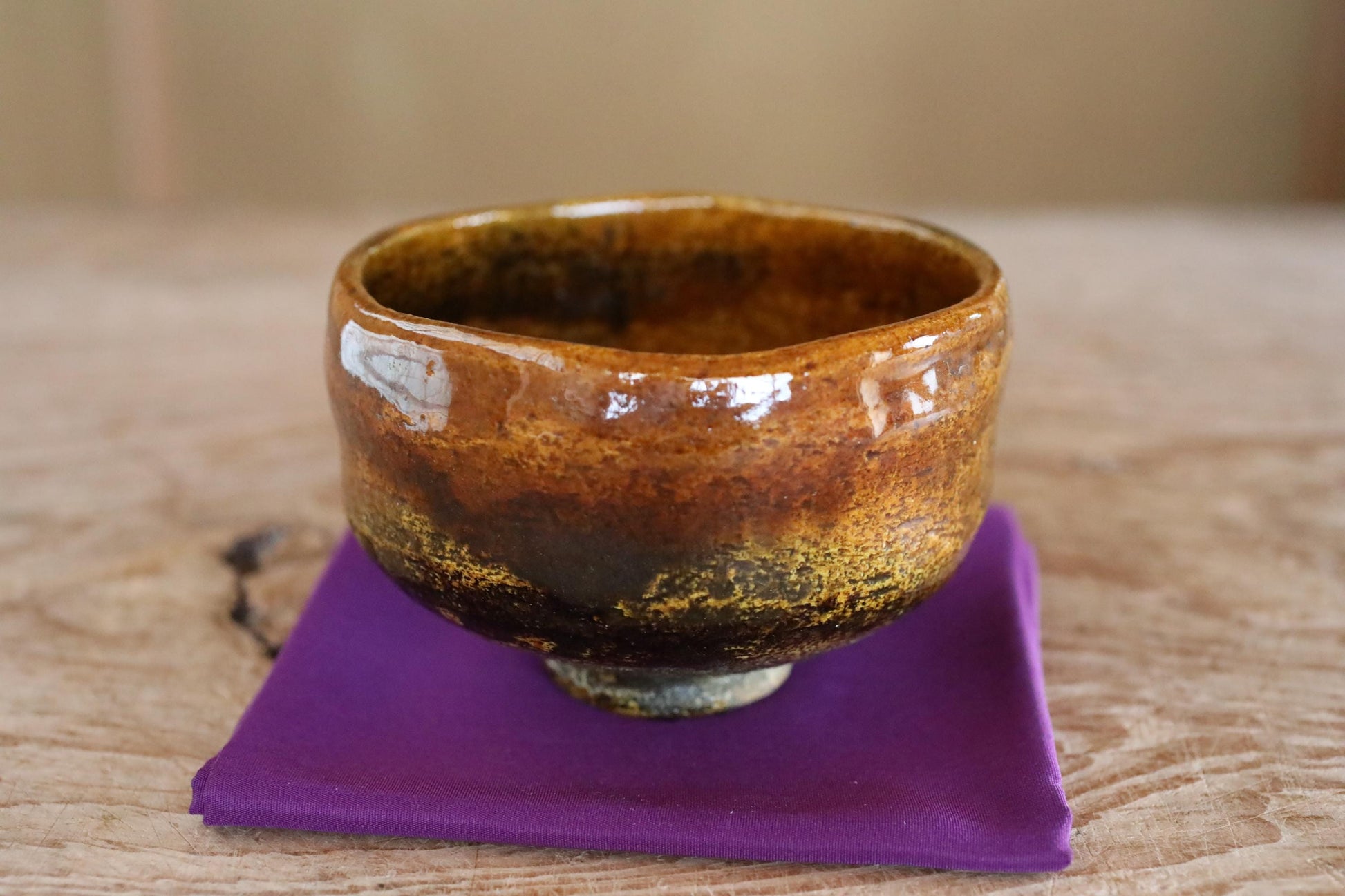 A ceramic bowl with a brown and orange glaze sits on a purple cloth on a wooden surface.
