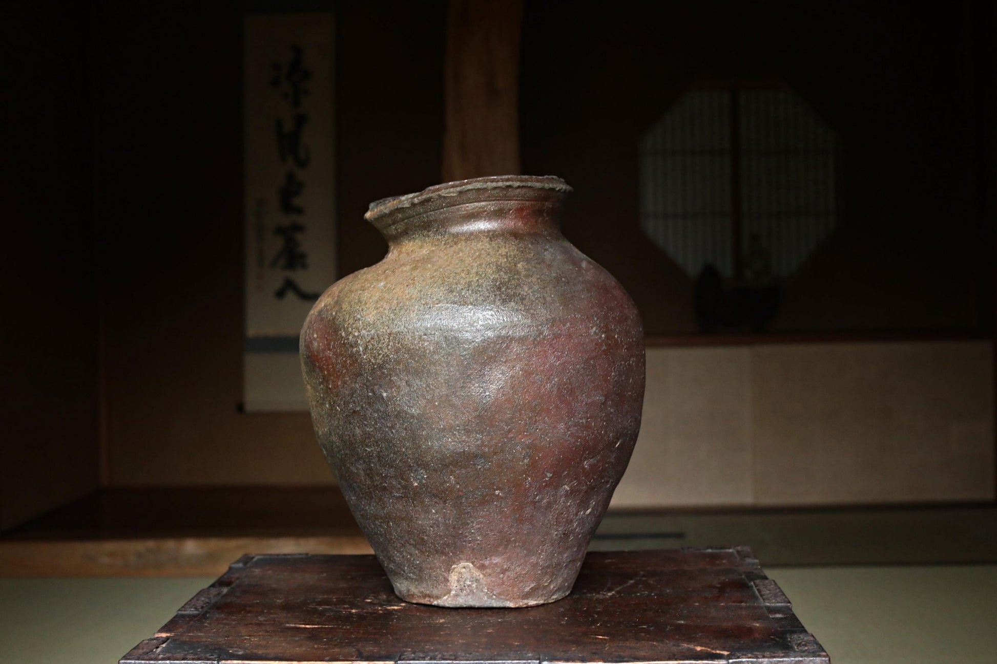 An old, weathered clay vase sits on a wooden surface, with a traditional Japanese-style window in the background.