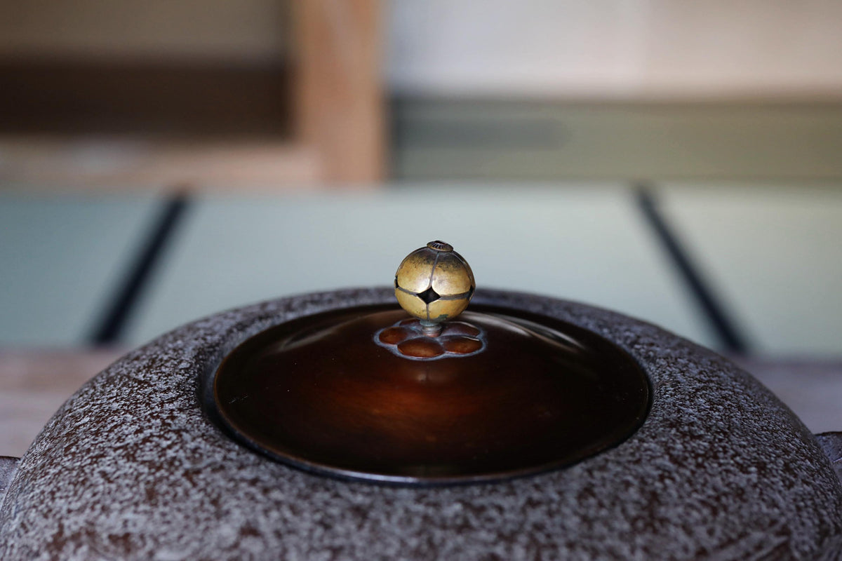 A close-up image of a dark brown teapot lid with a small bell-shaped object on top, placed on a wooden surface.