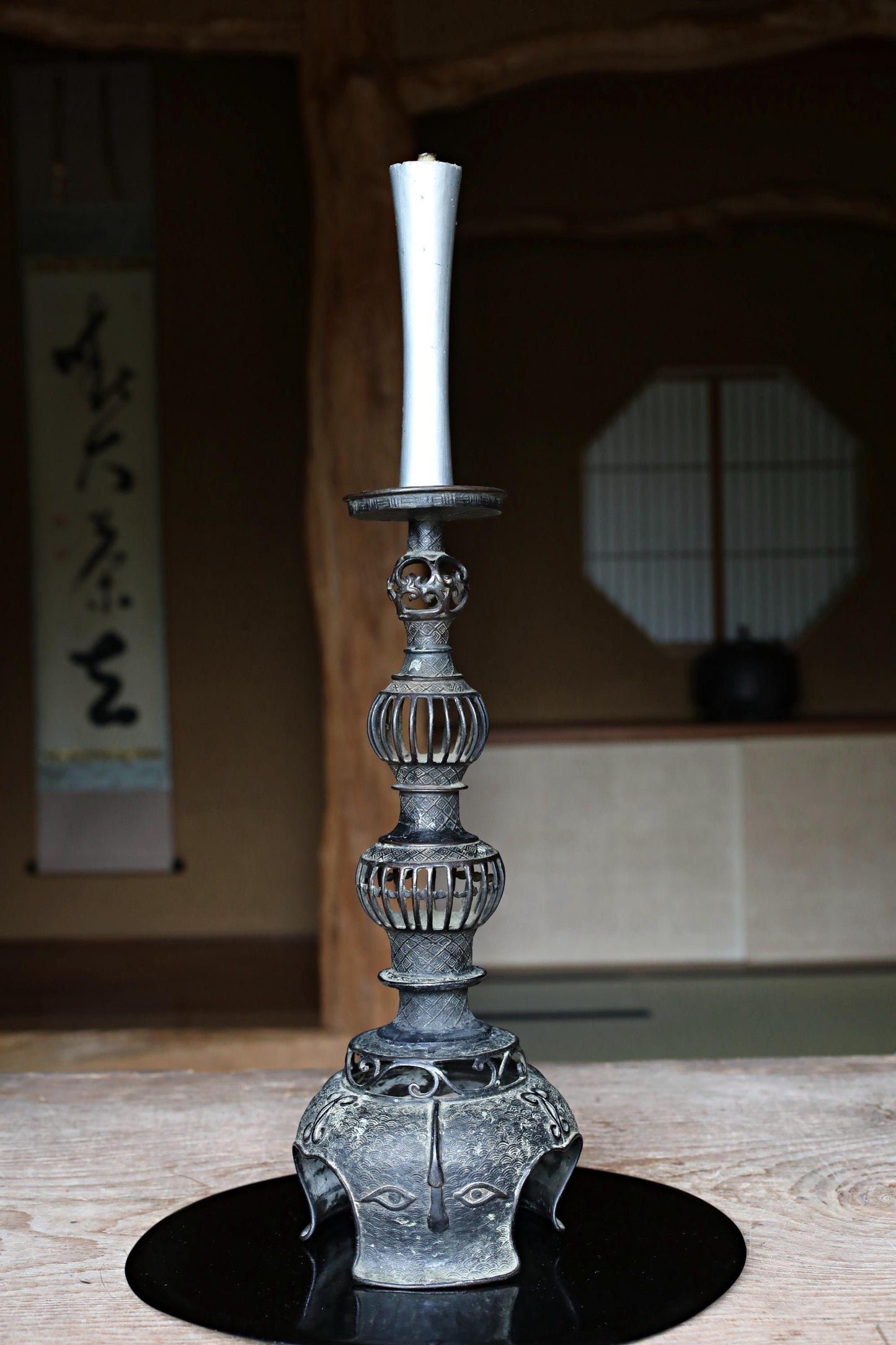 A decorative metal candlestick with a tall, slender white candle stands on a black circular base, placed on a wooden surface.
