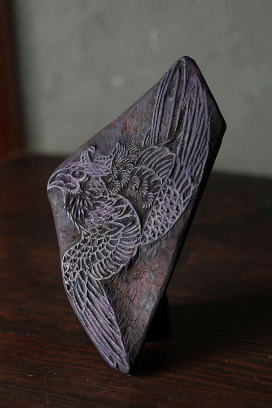 A dark purple, ornately carved wooden block or sculpture depicting a bird in flight.