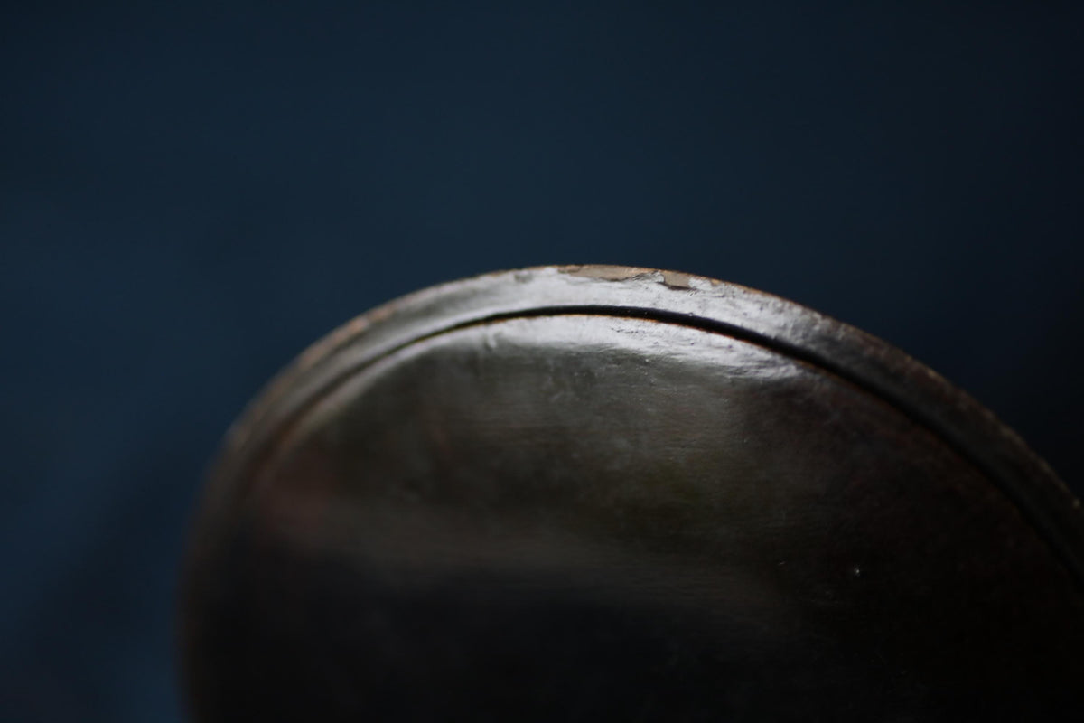 A close-up view of a dark, shiny surface, possibly a metal or glass object, with a slight curve and a small crack or mark visible.