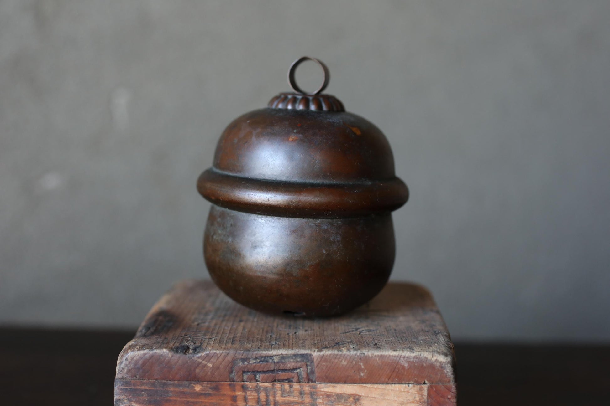 A round, metal object with a decorative top sits on a wooden surface against a gray background.