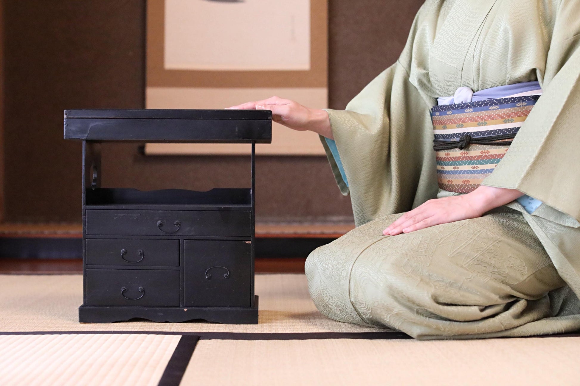 A person in traditional Japanese attire, including a kimono, is kneeling on a tatami mat and touching a small black wooden cabinet.