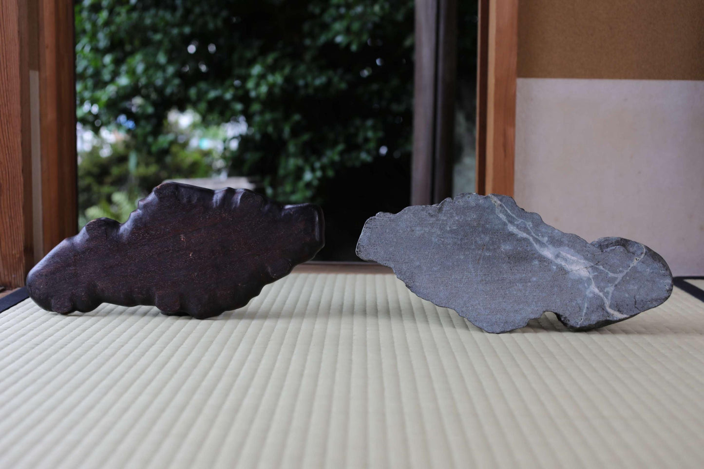 Two large rocks, one dark and one gray, are placed on a white surface, with a blurred background of trees.