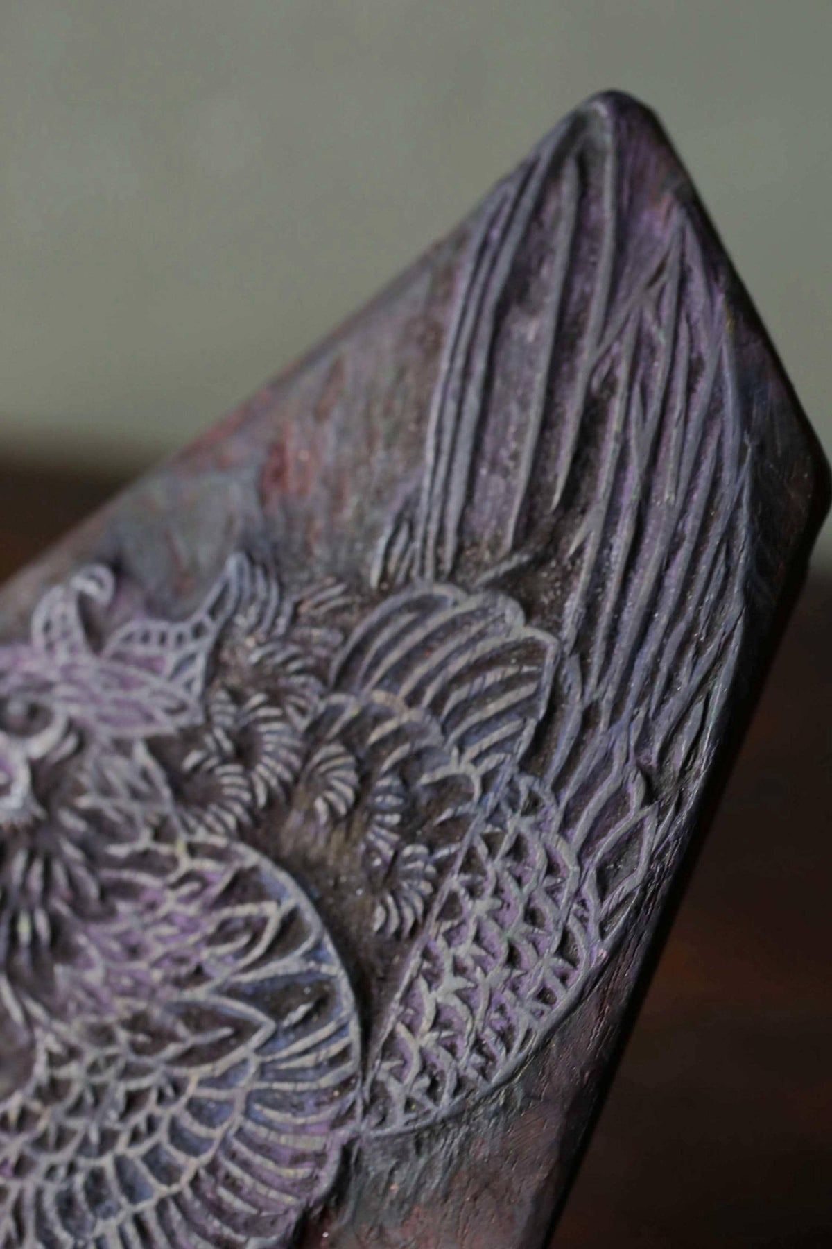 a close-up view of a detailed, intricately carved wooden object, possibly a piece of furniture or a decorative item. The object has a dark, textured surface with a pattern that resembles the feathers of a bird or a similar natural element. The image does not contain any text or other discernible objects or scenes.