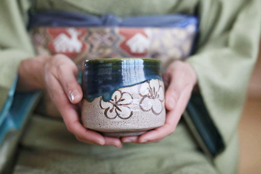 A person wearing a green kimono holds a ceramic mug with a floral design.