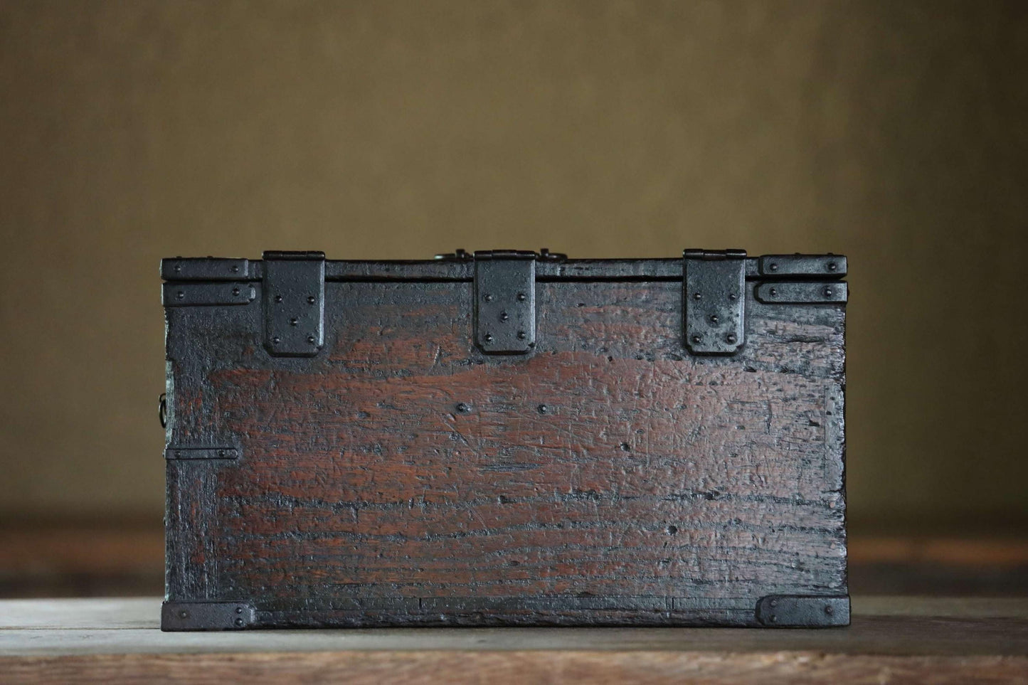 A weathered, wooden chest with metal clasps sits on a wooden surface against a plain background.