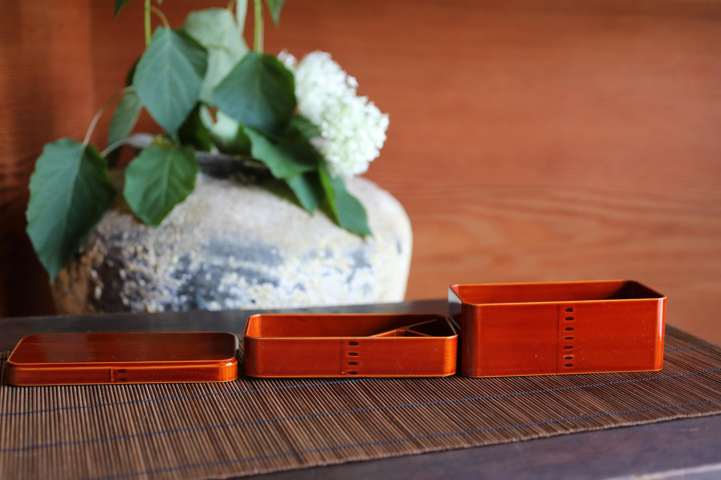 three orange rectangular containers placed on a wooden surface, with a vase of white flowers in the background.