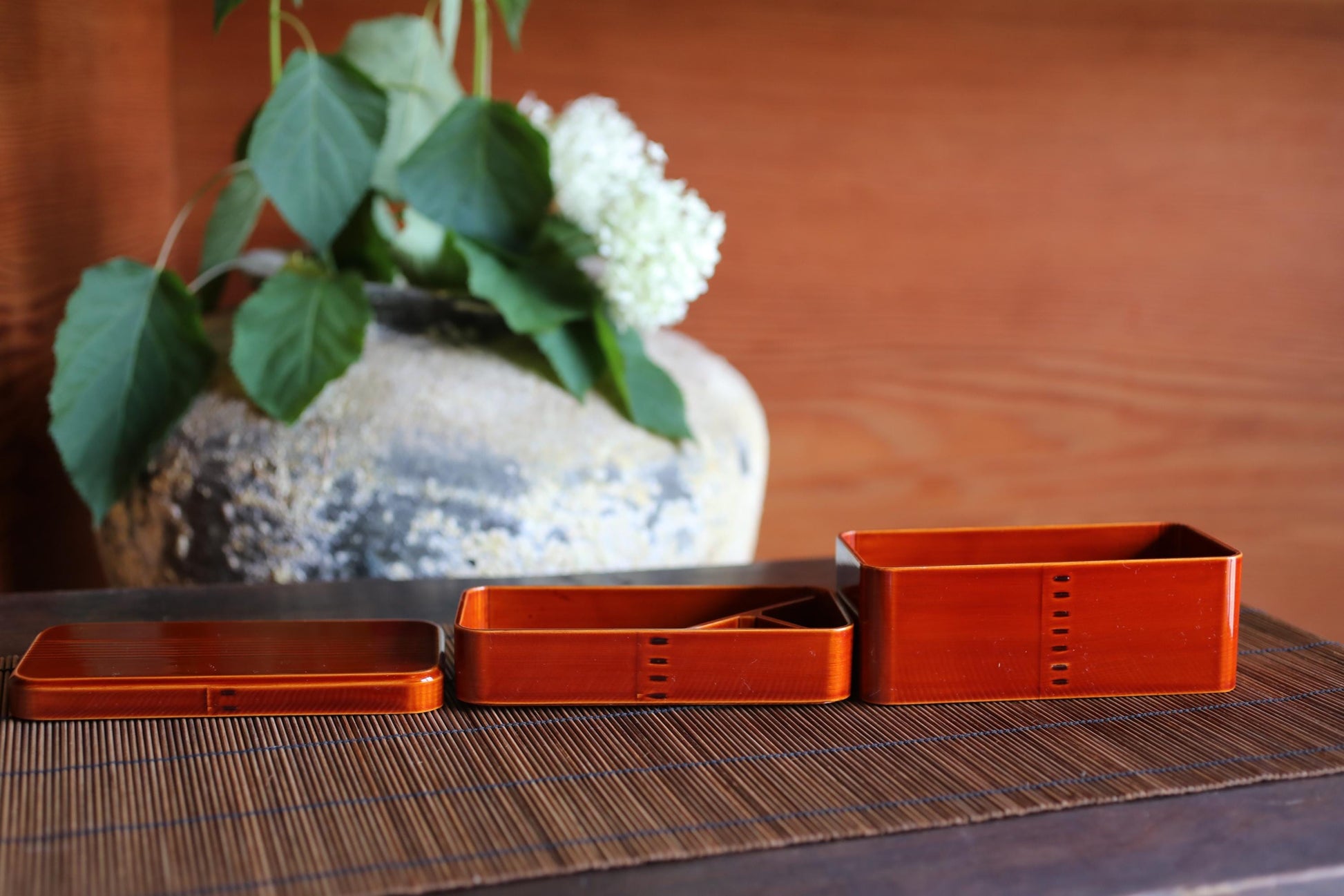 three orange rectangular containers placed on a wooden surface, with a vase of white flowers in the background.