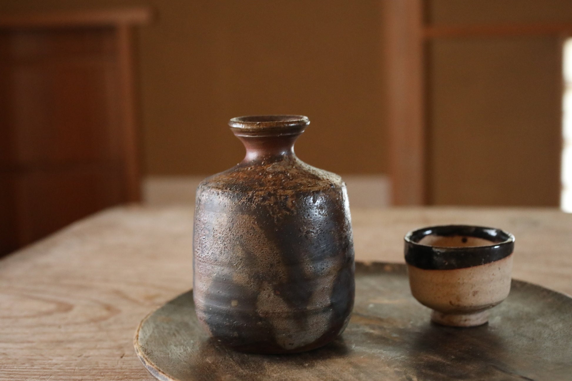 以下が含まれることがあります： 濃い釉薬がかかった茶色の陶器の酒瓶と、木製のトレイの上にある小さな白い陶器の酒杯