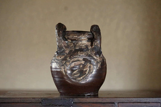 An old, weathered vase with a dark brown color and a distinctive design sits on a wooden surface against a plain background.