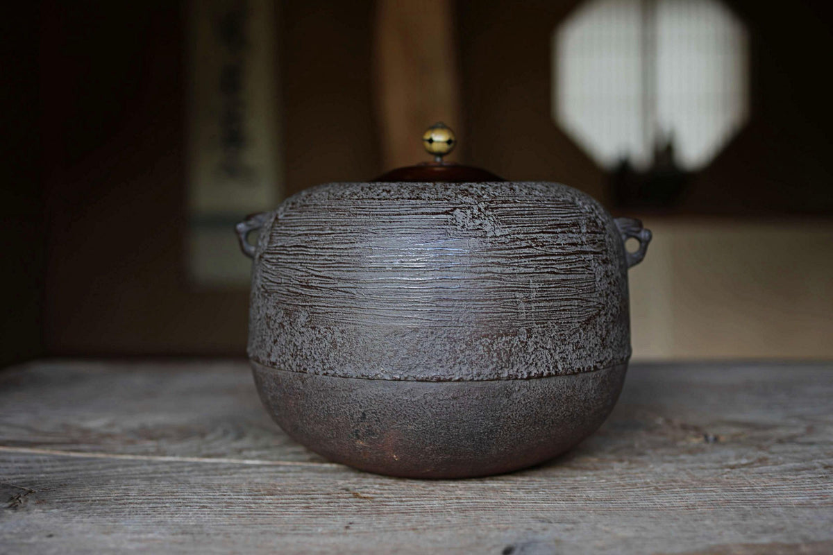 A dark-colored ceramic teapot with a gold-colored lid sits on a wooden surface, with a blurred background suggesting an indoor setting.