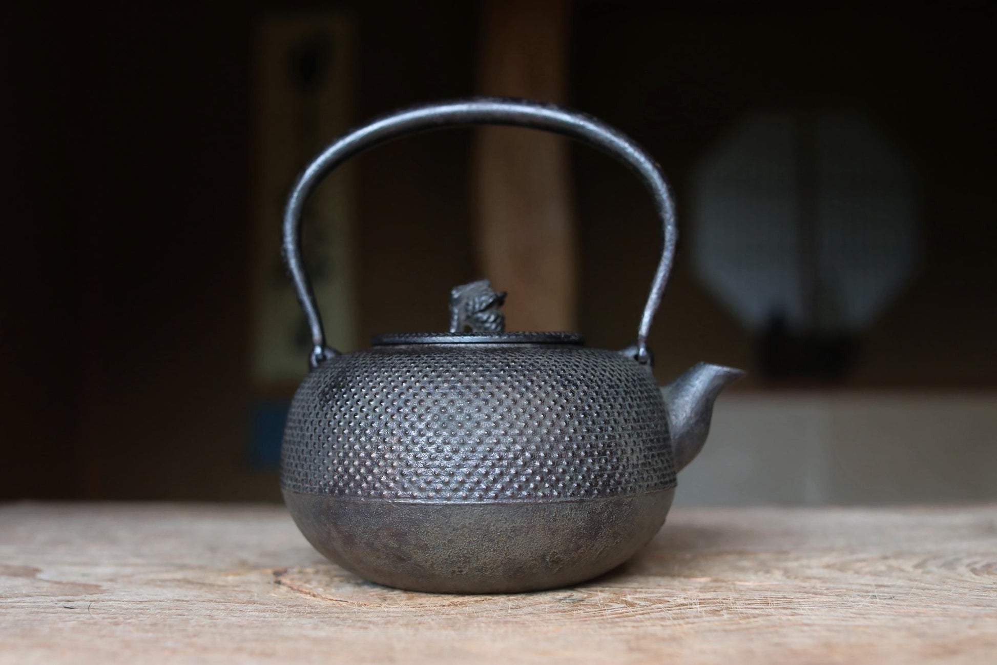 A black metal teapot with a dragon design sits on a wooden surface, with a blurred background suggesting a traditional Japanese setting.