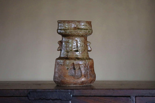 An old, weathered ceramic vase with a unique, textured design sits on a wooden surface against a plain background.