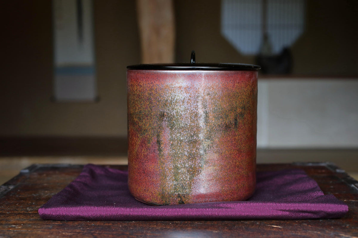 A ceramic container with a red and green speckled pattern sits on a purple cloth on a wooden surface.