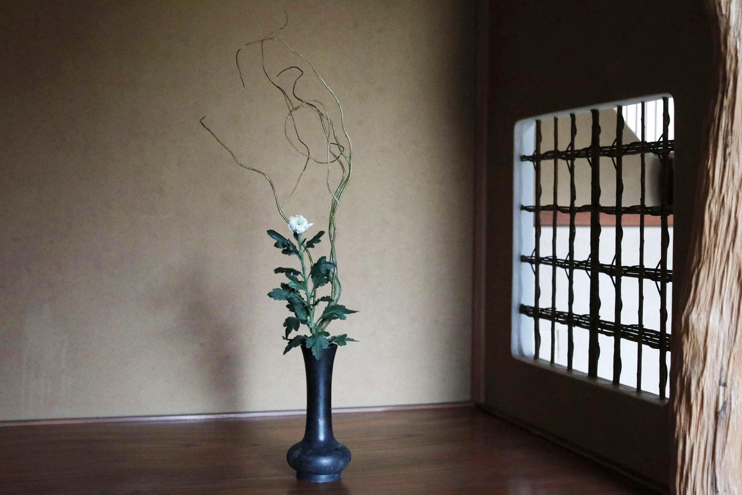 以下が含まれることがあります： 白い花、緑の葉、細い枝が入った黒い花瓶。花瓶は暗い木製の表面に置かれ、背景には窓と壁があります。