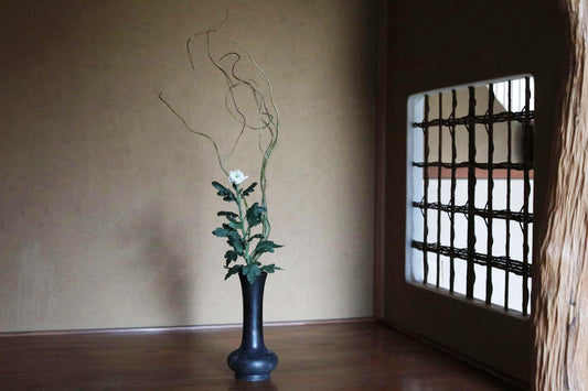 以下が含まれることがあります： 白い花、緑の葉、細い枝が入った黒い花瓶。花瓶は暗い木製の表面に置かれ、背景には窓と壁があります。