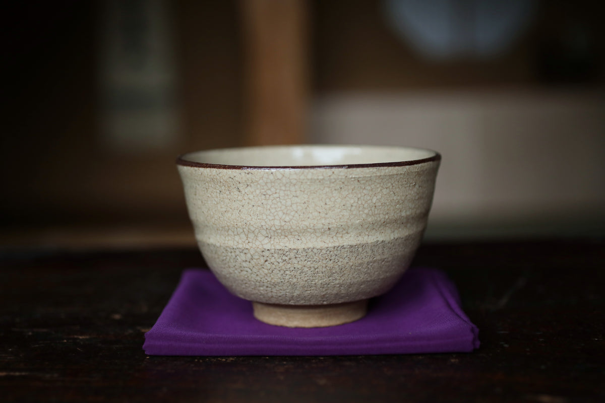 A white ceramic bowl with a brown rim sits on a purple cloth on a dark wooden surface.