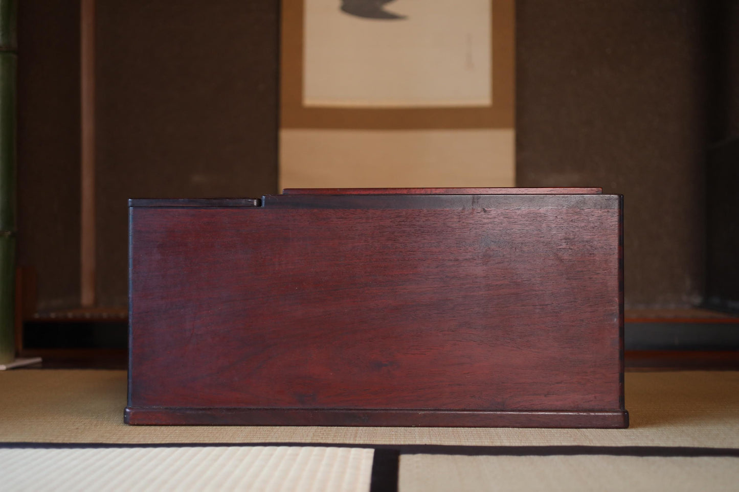 A dark wooden box sits on a tatami mat in a room with a traditional Japanese-style wall.