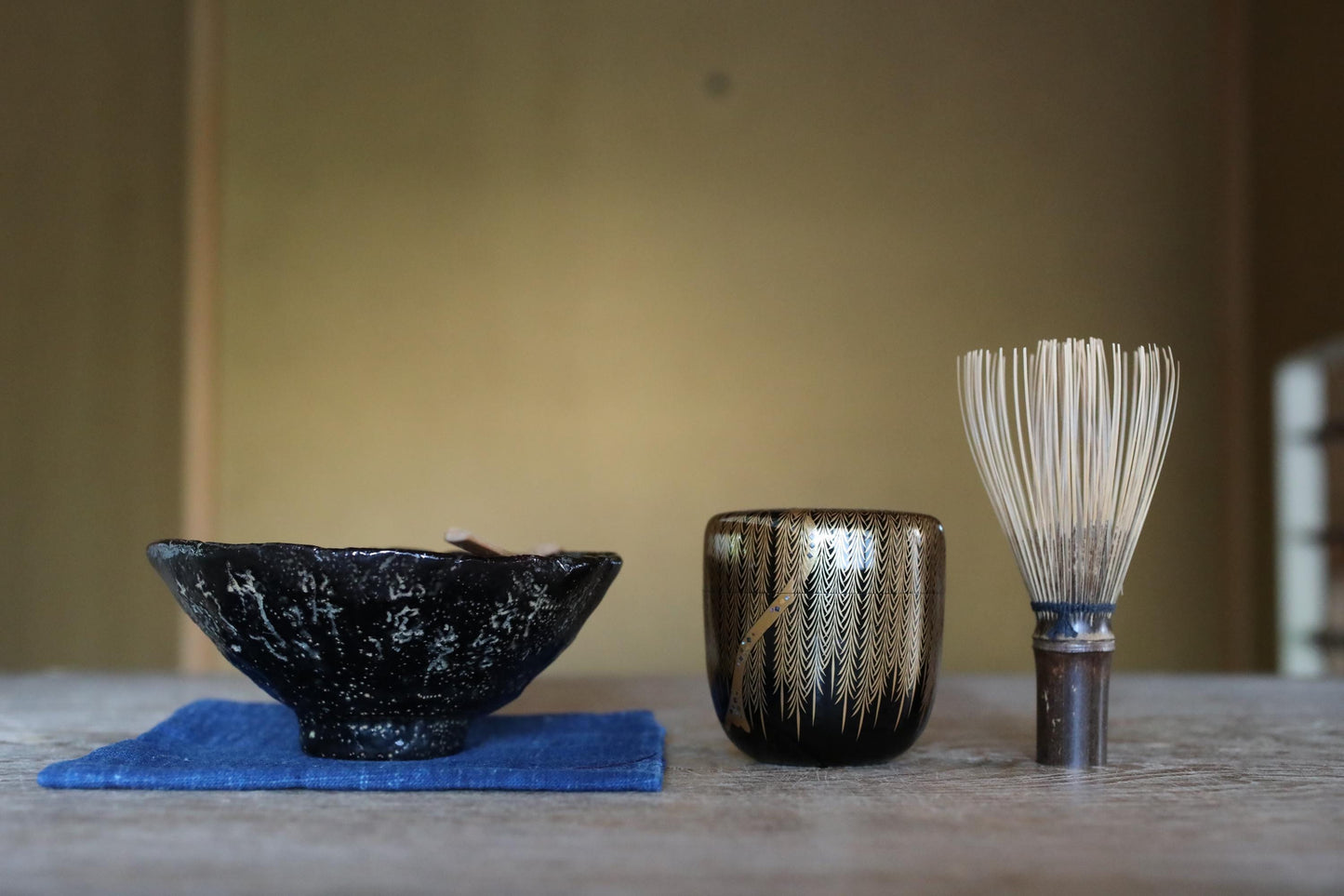 a black bowl, a black and gold cup, and a white whisk on a wooden table, all placed on a blue cloth.