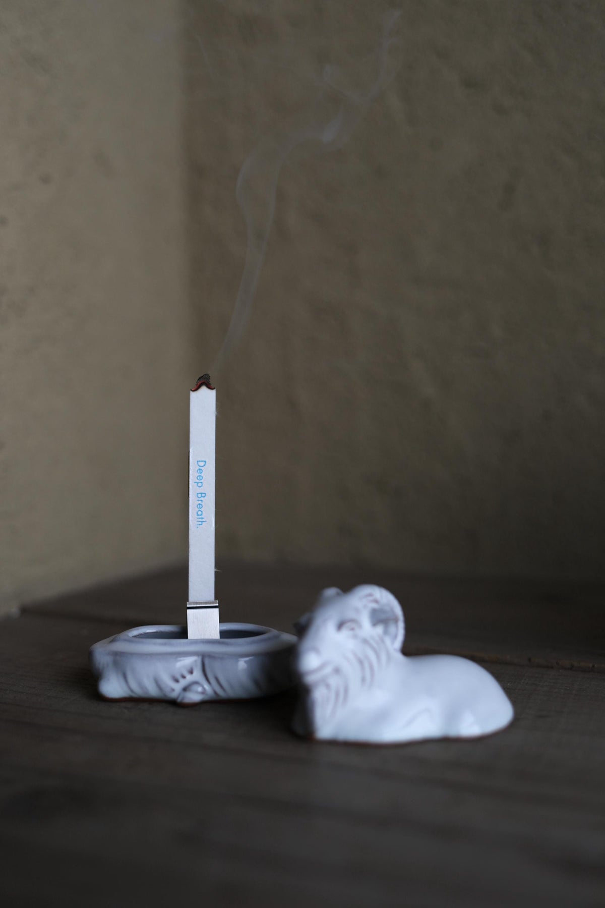 A white ceramic incense holder with a lit incense stick emitting smoke, placed on a wooden surface.