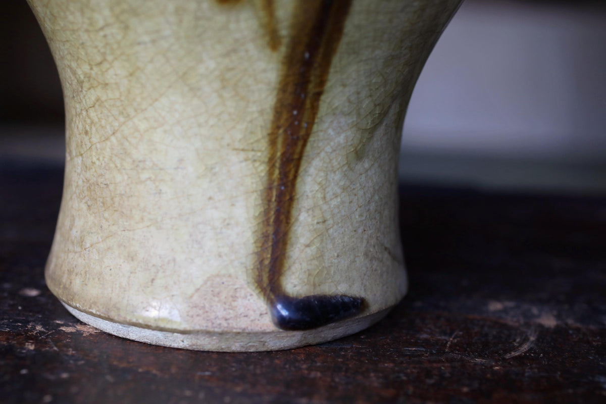 A close-up view of a ceramic vase with a dark brown drizzle or stain on its surface, resting on a dark brown surface.