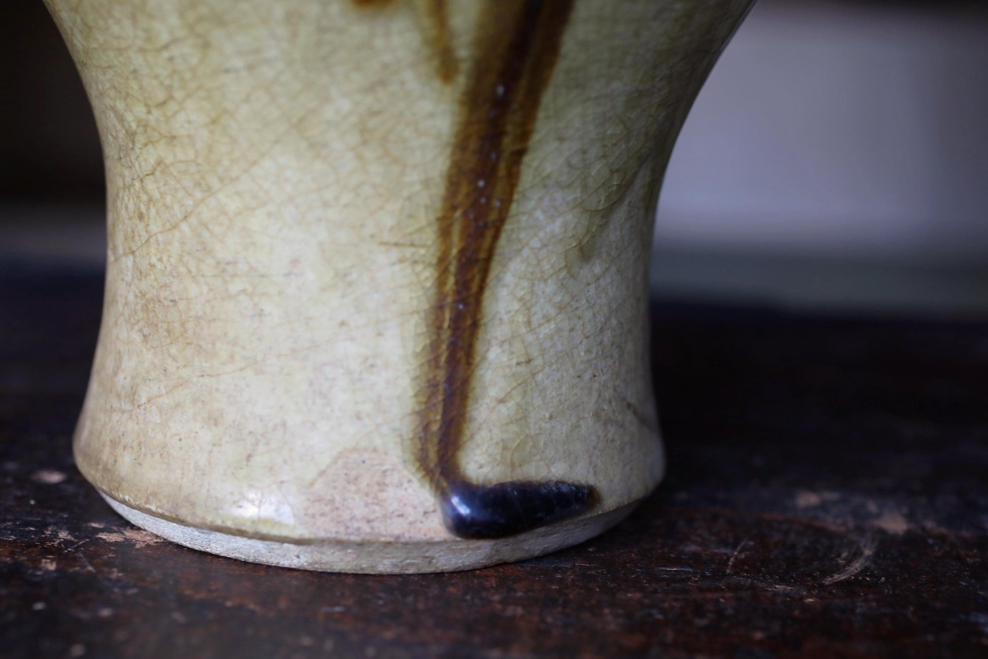 A close-up view of a ceramic vase with a dark brown drizzle or stain on its surface, resting on a dark brown surface.