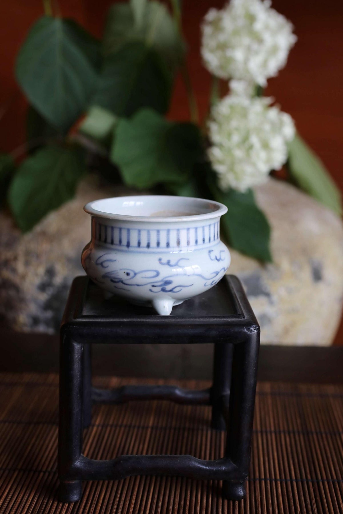 A white ceramic bowl with blue designs sits on a black metal stand, with a vase of white flowers in the background.