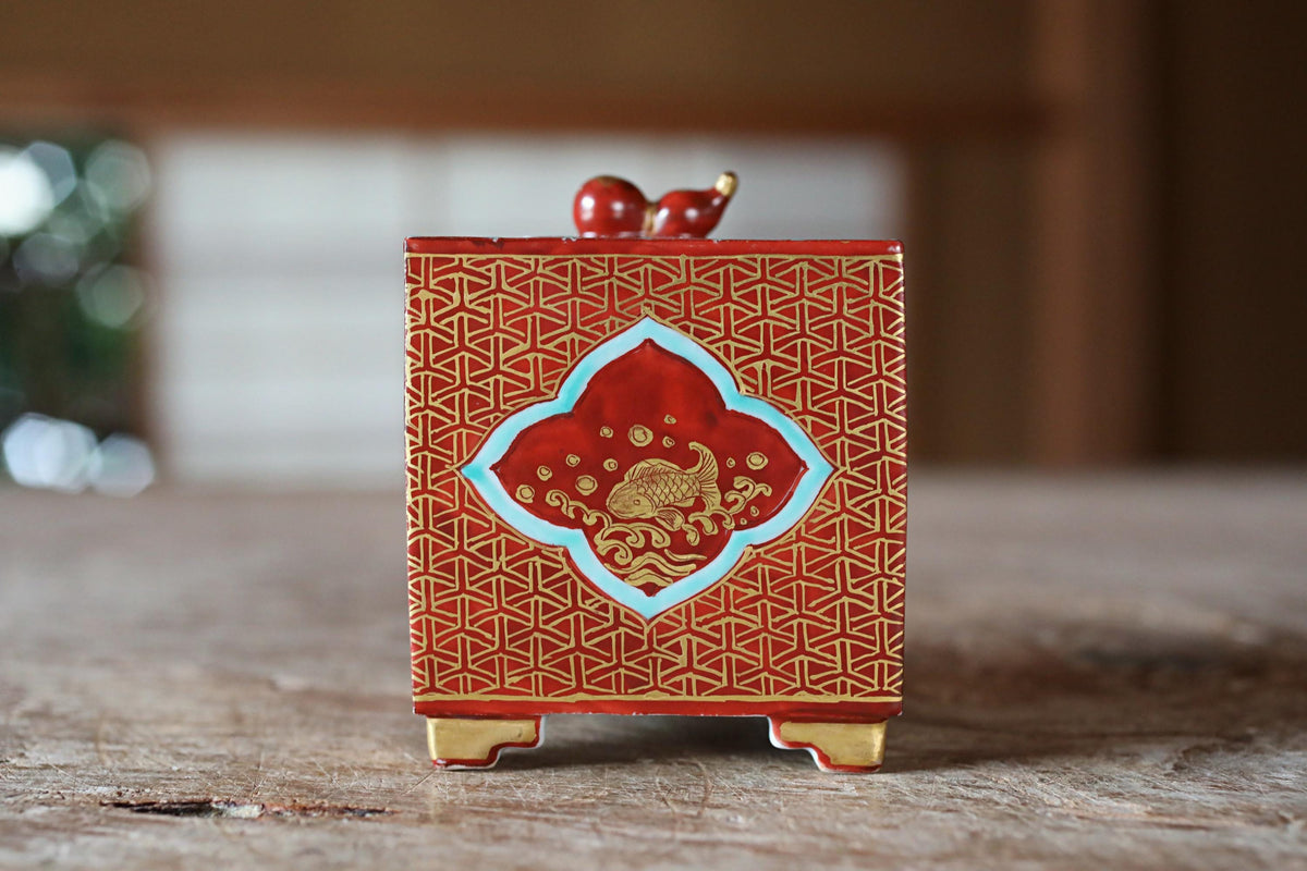 A red and gold decorative box with a patterned design and two small red figures on top, placed on a wooden surface.