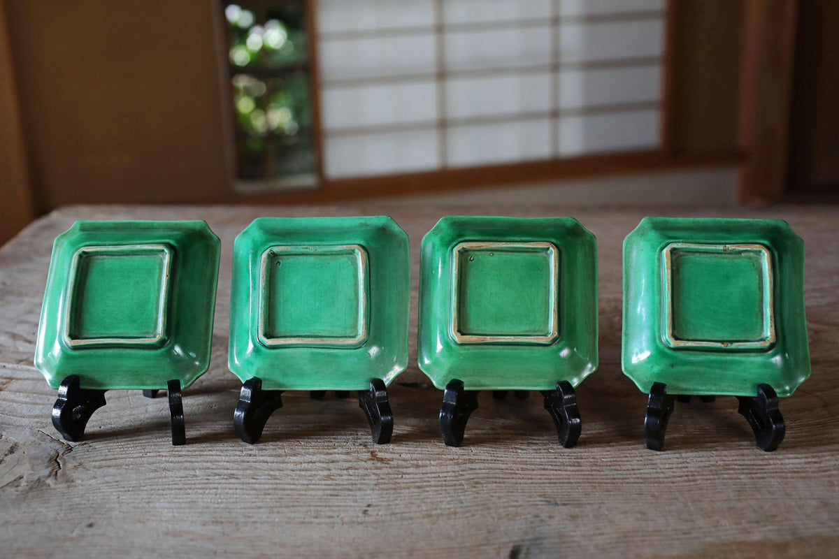 Four square green plates with gold trim are lined up on a wooden surface, each standing on a black stand.