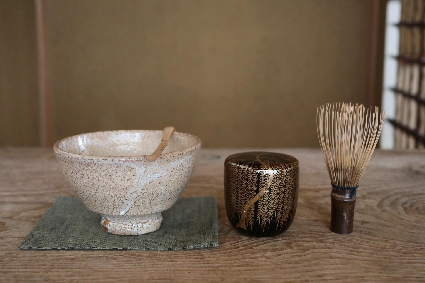 A white ceramic bowl with a wooden spoon, a black and gold ceramic container, and a wooden whisk are arranged on a wooden surface.