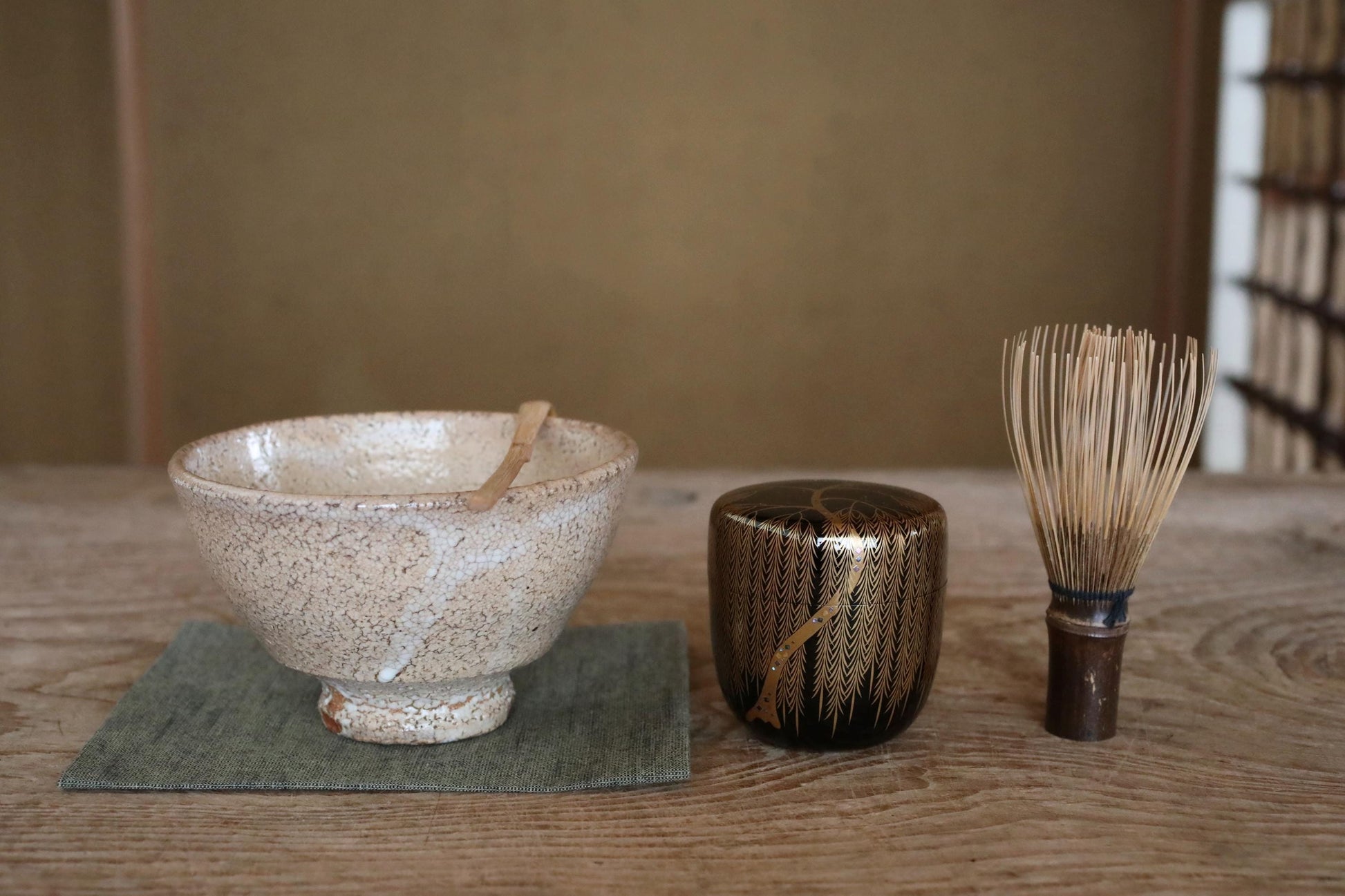 A white ceramic bowl with a wooden spoon, a black and gold ceramic container, and a wooden whisk are arranged on a wooden surface.