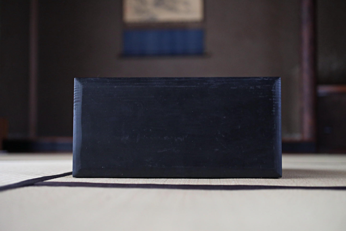 A black rectangular object is placed on a white surface, with a blurred background that includes a wooden structure and a blue and white patterned wall.
