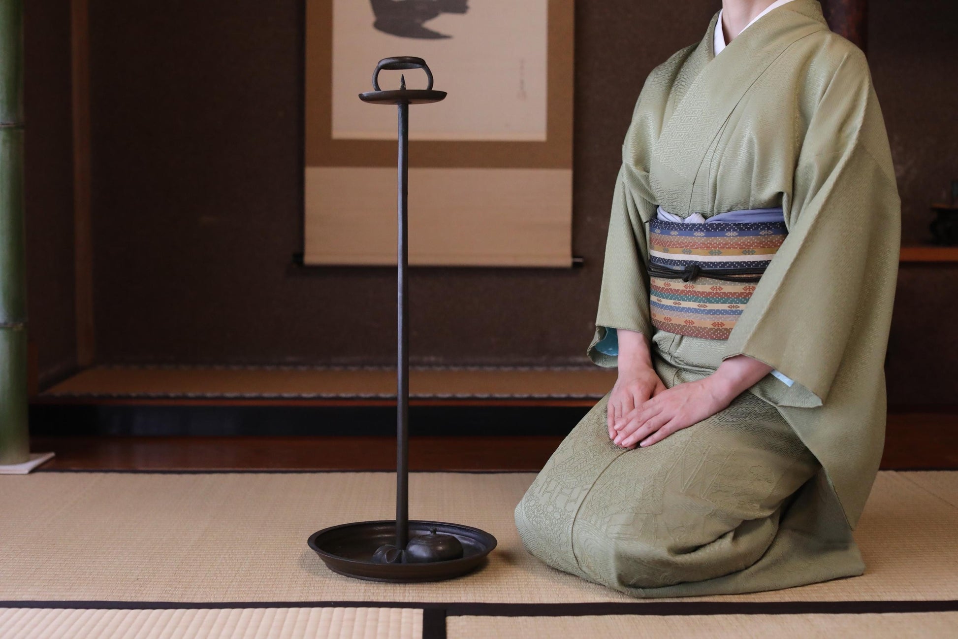 A person in a traditional Japanese kimono is kneeling on a tatami mat, with a small black object, possibly a tea set, placed next to them.