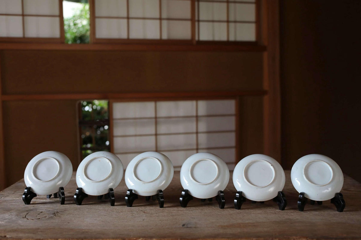 A row of six white plates or saucers lined up on a wooden surface, with a traditional Japanese-style building visible in the background.