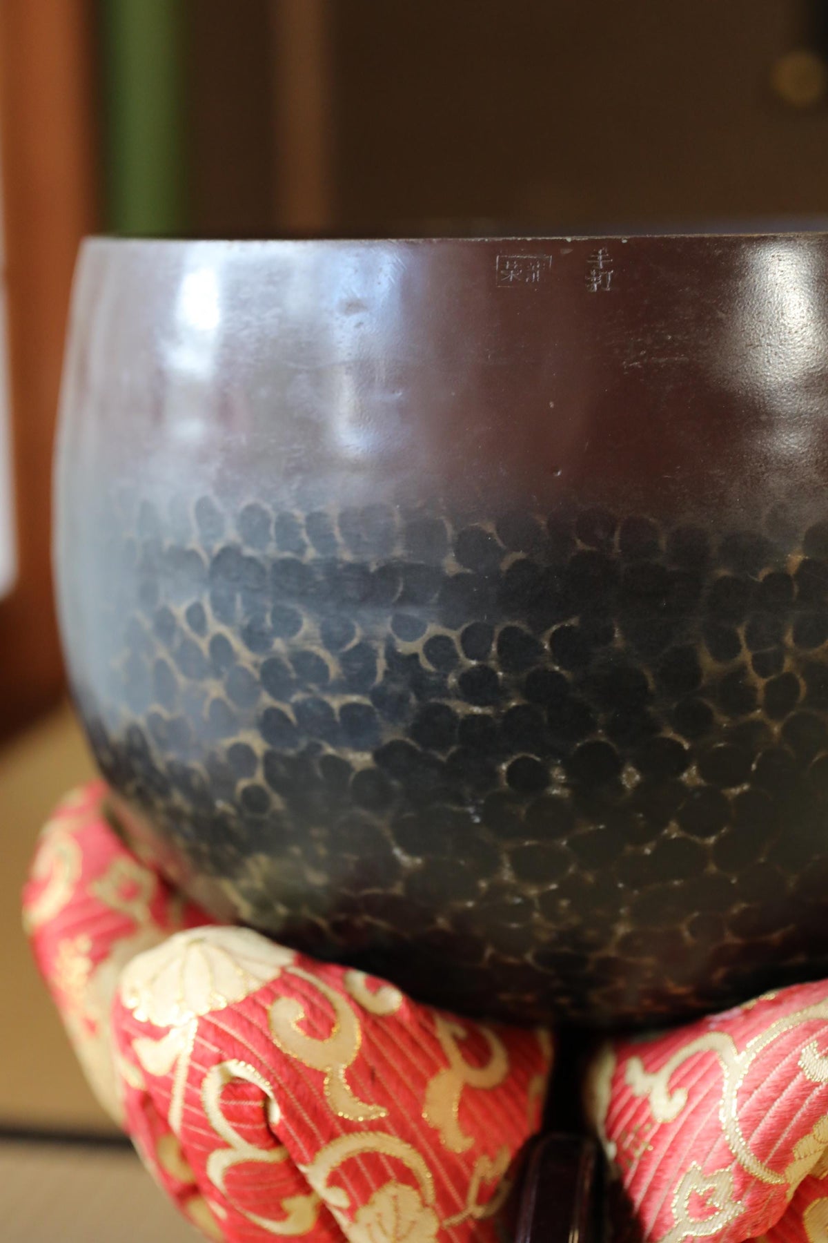 A close-up view of a dark-colored bowl with a textured surface, resting on a red and gold patterned cloth.