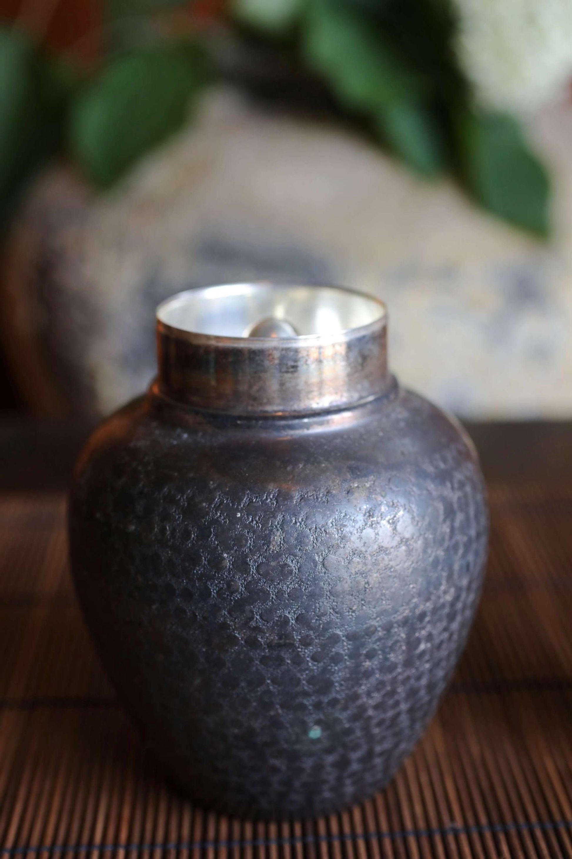 A dark-colored metal jar with a silver lid sits on a wooden surface, with a blurred background of green leaves.