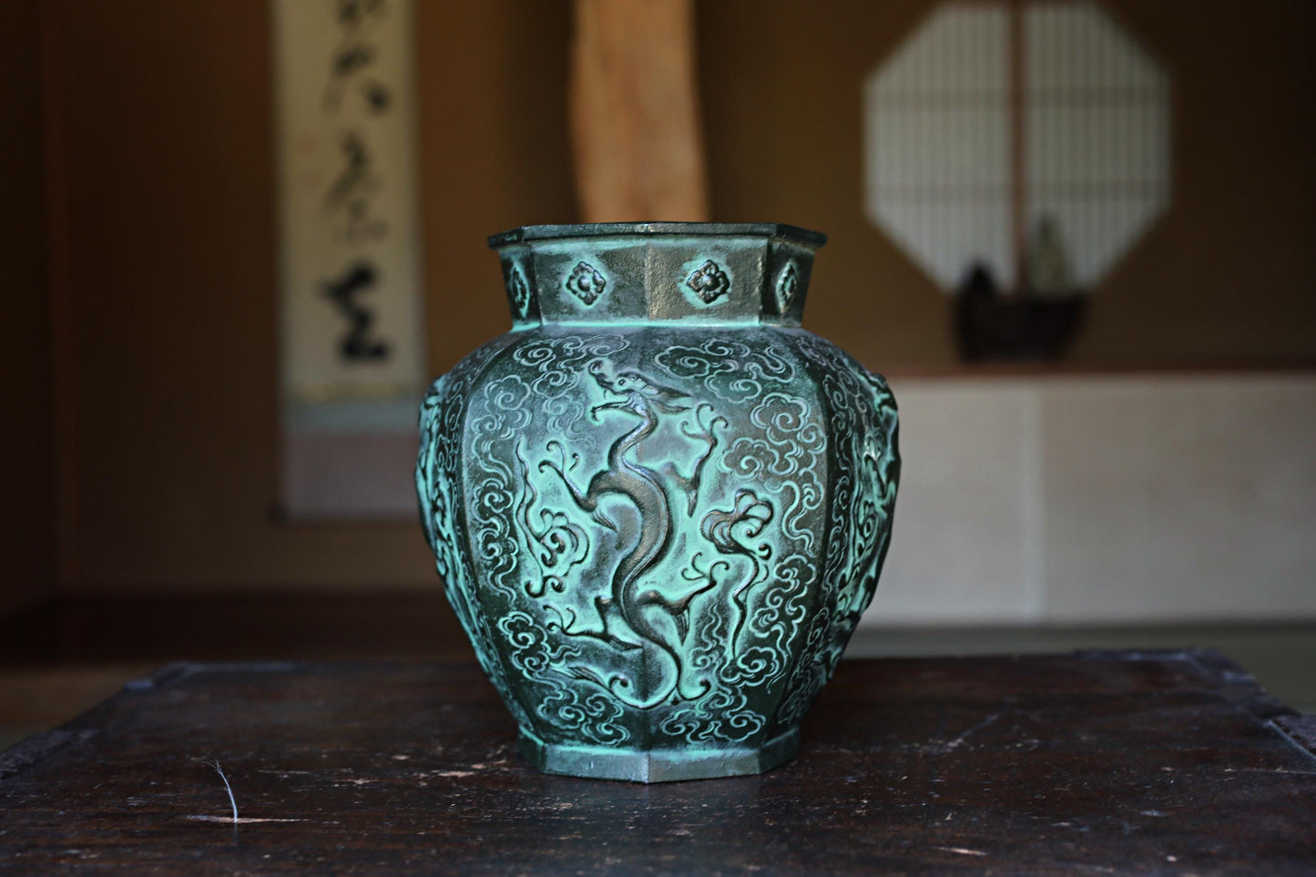 A decorative green vase with intricate patterns sits on a wooden surface, with a blurred background that includes a window and a wall with a traditional Japanese design.